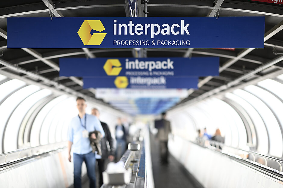 In einer tunnelartigen Struktur laufen die Menschen auf einem Förderband unter mehreren blauen Schildern mit dem gelben Interpack-Logo und dem Text „interpack PROCESSING & PACKAGING“ hindurch.